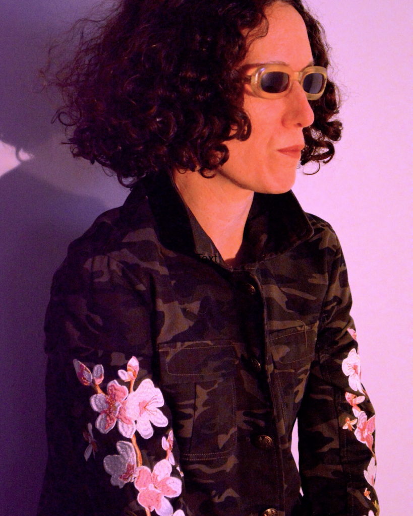 Side view of Analog Tara, against a lavender background, wearing vintage sunglasses and a green camoflouge jacket with embroidered pink and orange flowers on both sleeves.